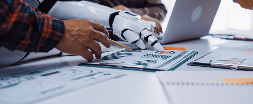 A modern workplace where a human hand collaborates with a robotic arm to analyze charts and business metrics, showcasing the integration of technology and teamwork.Noogenesis - Powered by Adobe