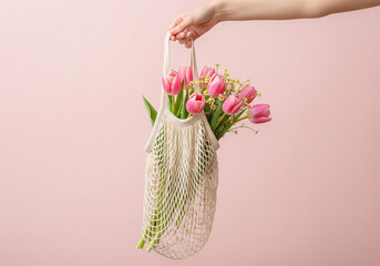 A hand holds a reusable mesh tote bag filled with a beautiful bouquet of pink tulips and mimosa flowers against a soft pink background.