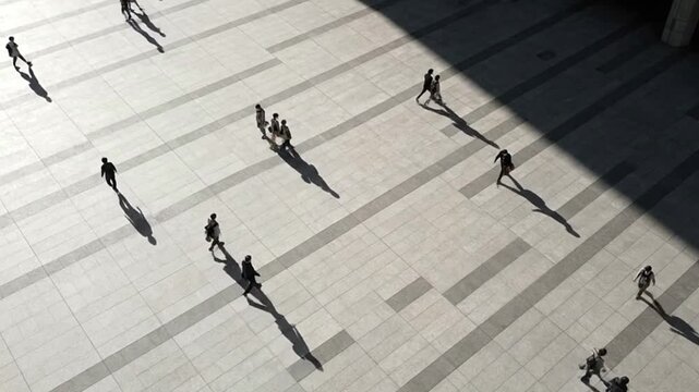 Highangle view of people with long shadows walking on a light gray paved plaza on a sunny day