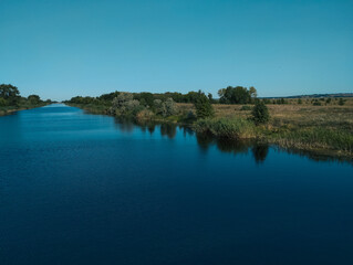 A calm, wide river with dark water separates green shrubs on the left bank and an open field on the right. A clear, cloudless sky complements the landscape.