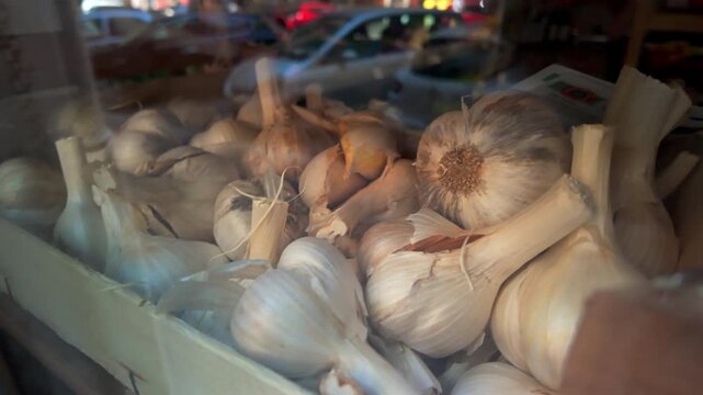 Wooden crate filled with dried garlic bulbs in a market display with reflections of passing cars and urban street life in the window glass