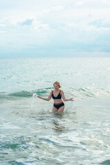 A woman in a bikini stands in the ocean, splashing water, surrounded by soft waves under a cloudy sky.