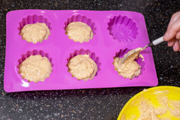 A hand carefully spoons batter into a silicone baking pan, preparing individual servings of banana muffins.