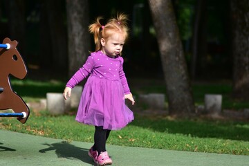 Fototapeta premium Red-haired toddler in purple dress and pink sandals standing on playground trampoline, looking down with curiosity, blurred horse spring rider behind, green park surroundings.