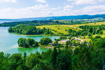 A turquoise lake surrounded by green meadows and trees, with roads and houses visible near Czorsztyn Castle under a blue summer sky. The scene showcases Polish nature with calm waters, rolling hills, 