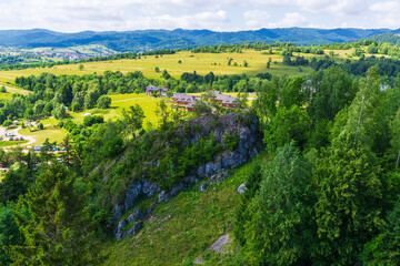 Obraz premium Rolling green hills and scattered houses with rocky cliffs in the foreground, viewed from the area near Czorsztyn Castle on a sunny summer day. This rural mountain landscape in Poland features lush 
