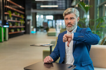 Dislike. Upset businessman guy in suit sitting in office lobby showing thumb down sign gesture, expressing discontent, disapproval, dissatisfied bad work. Displeased serious mature man in hotel hall.