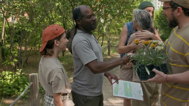 Medium shot of joyous young Caucasian family with young son greeting senior diverse neighbor couple, shaking hands and hugging, before planting flowers together in community garden