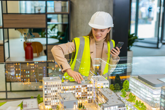 Confident female girl architect in hardhat and reflective jacket using smartphone and analyzing business office complex architectural model. Woman lady smart designer of buildings working in office. - Powered by Adobe
