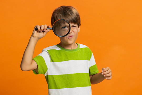 Investigator researcher scientist preteen child boy kid holding magnifying glass near face, looking into camera with big zoomed funny eye, searching, analyzing. Young children on orange background