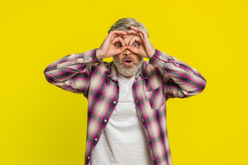 Mature man forms binocular shape with hands, peering into distance and sparking humorous detective flair. Middle-aged guy on yellow background anticipation expecting looking for a friend waving hello