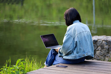 Woman in jeans jacket sitting with laptop and  smartphone on lake beach. Concept of remote work outdoors at summer park, girl freelancer