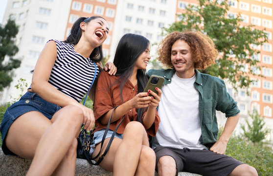 Three cheerful multi-ethnic young friends are sitting together outdoors, enjoying their time together while using a smartphone - Powered by Adobe