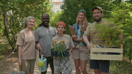 Medium full portrait of active multiethnic neighbors, holding tree saplings, flower plugs, gardening tools, standing in local park on summer day, looking at camera, smiling with joy