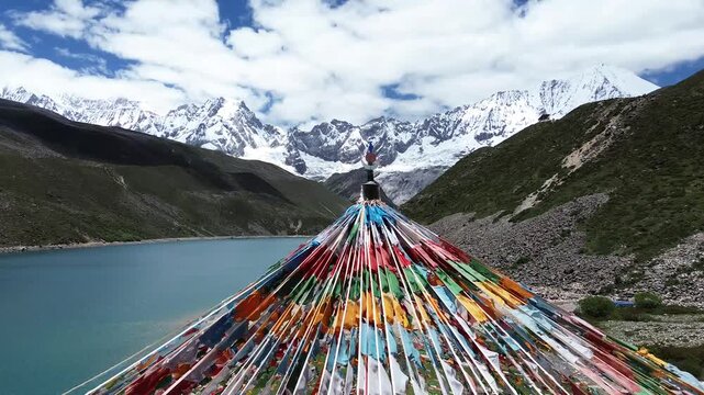 Drone shot gliding over vibrant Tibetan prayer flags, unveiling Pemaling Lake with the majestic Kula Kangri Massifs in Lhoka, Shannan, Tibet, in the heart of the Tibetan Himalayas.