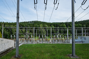 Front view of electrical substation with high voltage lines, insulators, and transformers, surrounded by green grass and forest under blue sky.