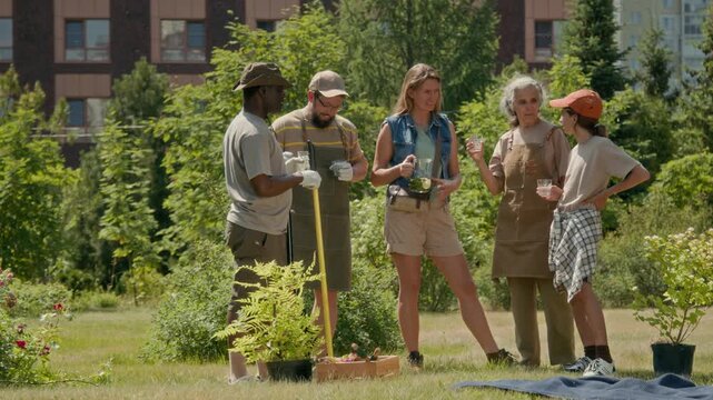 Full shot of happy, friendly diverse group of neighbors living in urban apartment complex working together in community garden in summer, drinking lemonade and chatting while taking rest break
