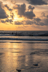 Distant silhouettes of three fishermen wading into the Arabian Sea at Caranzalem Beach, Goa, on a monsoon evening, with golden sunset, dramatic clouds, and shimmering water reflections.