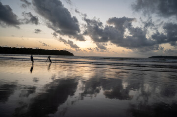 Dusk at Caranzalem Beach, Goa, with dramatic cloudy skies, waves, and two backlit cliffs. Two fishermen silhouettes cast their lines, their reflections mirrored in the wet sand and sea.

