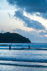 Two fishermen silhouettes fishing in the Arabian Sea at dusk, with a backlit cliff, orange glow, and grey monsoon clouds in the sky, shot from Miramar Beach, Panjim, Goa.

