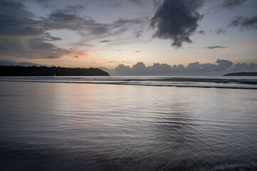 Obraz premium Landscape view of Miramar Beach Panjim Goa during monsoon sunset, with Arabian Sea waves, dramatic grey clouds, yellow-orange horizon glow, backlit cliffs, wet sand textures, and shimmering reflection