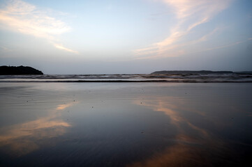 A shot of heart-shaped cirrus white clouds in the light blue sky at Miramar Beach, Goa, reflected in the water, with gentle waves and two distant cliffs in the background.