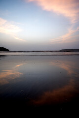 A shot of heart-shaped cirrus white clouds in the light blue sky at Miramar Beach, Goa, reflected in the water, with gentle waves and two distant cliffs in the background.
