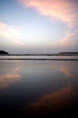 A shot of heart-shaped cirrus white clouds in the light blue sky at Miramar Beach, Goa, reflected in the water, with gentle waves and two distant cliffs in the background.