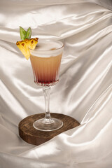 Red gradient cranberry cocktail with pineapple garnish served in a glass on a round wooden board, set against a clean white background.