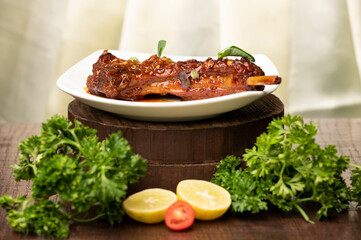 Lamb chops on white plate over wooden round board, styled with lemon slices, cherry tomatoes, coriander leaves in foreground, yellow satin background.
