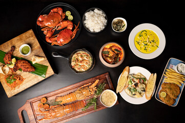 A top shot of assorted Indian and continental dishes on a black table including tandoori chicken, lobster, crab butter garlic, prawns, curry, pasta, risotto, fried fish, and avocado sandwich.
