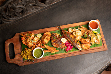 Top view of Goan seafood platter with rawa fried mackerel, pomfret, crab butter garlic, spiced panko prawns, and Sukhem Tisreo served with chutney on banana leaf in rustic setting.