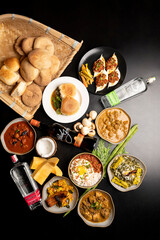  Top view of traditional Goan food spread on black table, including stuffed crabs, Ross omelette, poi and pao in wooden basket, chicken and mutton xacuti, pumpkin dish, and Feni bottles.