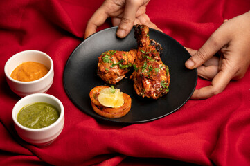 Juicy chicken tangdi kebab with fresh salad in black plate on red table mat background, green chutney and yellow sauce in white bowls, with hands placing the plate.