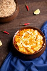 A top shot of Paneer Butter Masala, cottage cheese slow-cooked in rich buttery tomato and cashew nut gravy, served in a rustic brown pot on blue satin with green and red chilies.