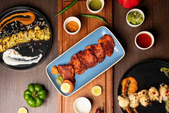 Top shot of Fish Tikka Masala, Murgh Malai Kebab, and Badami Broccoli styled with lemon, chutneys, red-green chillies on a brown platter over brown table.