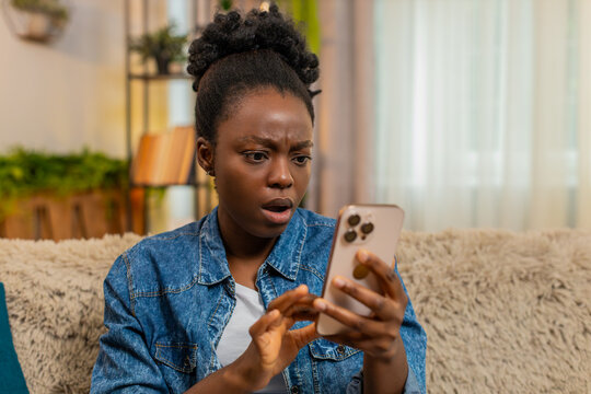 African American young woman suddenly opens her eyes wide in shock after reading unexpected and emotional social media news. Black girl stares at her phone, frozen by the intensity of what she sees.