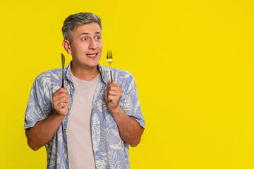 Middle-aged man holding knife and fork while looking directly at camera with eager hungry expression, showing desire for tasty meal. Young guy expressing appetite isolated on yellow background.