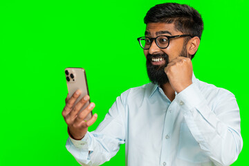 Indian man looks excited at smartphone screen, celebrating online game win, jackpot or great news with joy. Arabian guy isolated on green chroma key background showing victory reaction and happiness