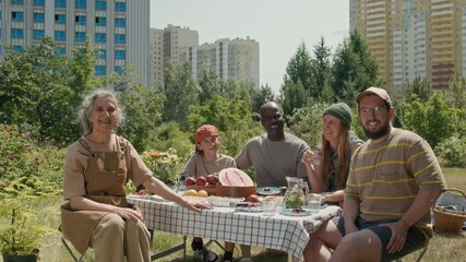 Medium portrait of group of diverse neighbors enjoying picnic after doing volunteer work, planting greenery in local garden. Young Caucasian family, African American husband and wife smiling at camera - Powered by Adobe