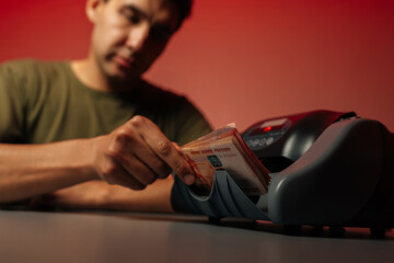 Close-up selective focus shot of serious cashier sitting at desk, counting cash with electronic money counter machine, studio shot on isolated red background. Concept of financial accounting.
