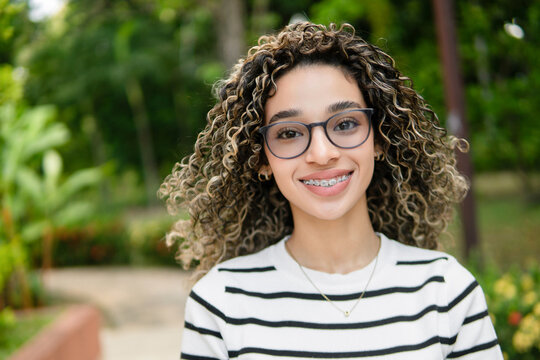 Portrait of a cheerful young hispanic university student smiling, wearing braces and glasses, in a park