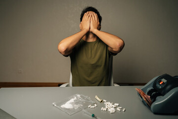 Studio shot of drug addiction struggle. Portrait of unrecognizable man covering face sitting at table with narcotics, banknotes, syringe, showing despair, dependency crisis. Concept of substance abuse