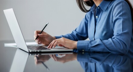 A woman in a blue shirt taking notes with a pencil next to a laptop on a reflective desk.