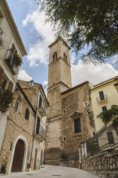 View of a tall, weathered stone tower rises above the ancient buildings, framed by the sky and foliage, casting shadows on narrow street, Pacentro, Abruzzo, Italy.