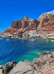 Obraz premium Picturesque harbor at the base of red volcanic cliffs, with boats floating on turquoise water in a charming Greek coastal village.