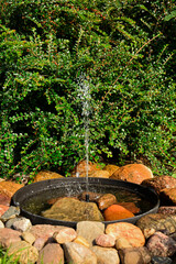 źródełko w ogrodzie, mini fontanna solarna otoczona kamieniami i zielonymi roślinami, tryskający strumień wody z małej fontanny ogrodowej, a solar fountain in a small bowl in the garden