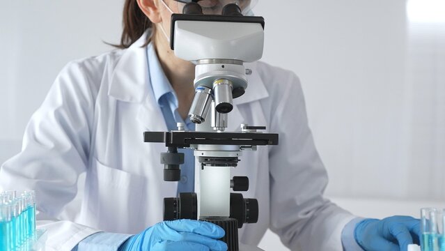 Female scientist wearing protective gear carefully adjusting microscope lens, analyzing medical research samples with precision in sterile laboratory environment