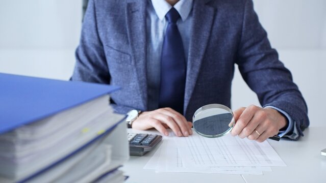 Professional businessman examining financial documents, magnifying glass in hand, calculator positioned on sleek desk workspace. Audit and taxes in business