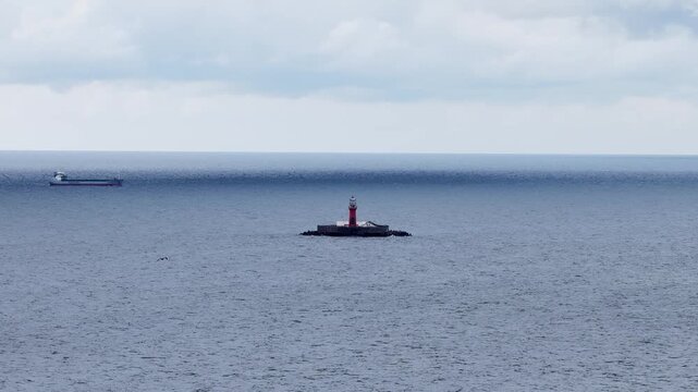 Distant view of a red offshore lighthouse on a small rocky island in the Baltic Sea near Kolka, Latvia, with a cargo ship sailing in the background under cloudy skies.